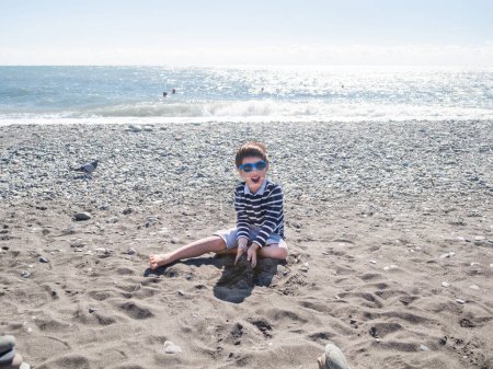 Téléchargez les photos : Happy boy construit une pyramide avec du sable mouillé, des pierres et du bois flotté sur une plage de galets. Vacances au bord de la mer. Explorer la nature dans l'enfance. - en image libre de droit
