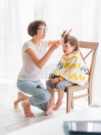 Téléchargez les photos : "Mère coupe les cheveux de son fils toute seule. Le petit garçon est assis, couvert de tissu, et tient une paire de ciseaux. Nouvelle normale en cas de quarantaine de coronavirus COVID-19. Beauté à la maison lifestyle." - en image libre de droit