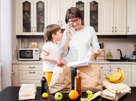 Photo for Woman and toddler boys sorts out purchases in the kitchen. Grocery delivery in paper bags. Online order from grocery store. Mother and son at kitchen. - Royalty Free Image