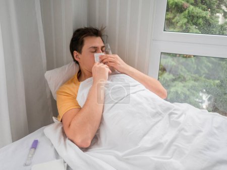 Photo for Sick man is lying in bed with thermometer. Man is blowing his nose. Temporary disability in case of disease symptoms. - Royalty Free Image