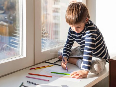 Foto de "El niño se sienta en el alféizar de la ventana y dibuja el arco iris con lápices de colores. Actividad creativa de ocio para los niños en casa." - Imagen libre de derechos