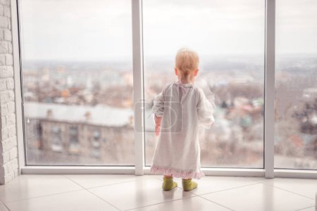 Photo for "little girl in a cute dress looks out the window" - Royalty Free Image