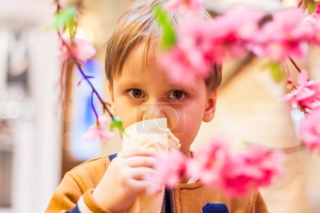 Téléchargez les photos : "Un enfant heureux mange de la glace. Un petit garçon souriant tient dans ses mains 1 glace, cachée par les branches fleuries d'un arbre." - en image libre de droit