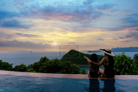 Téléchargez les photos : "Femmes asiatiques en maillot de bain noir avec sa fille assise sur le côté de la piscine à débordement relaxant au coucher du soleil avec vue sur la mer pendant la retraite de vacances. Famille heureux et profiter avec vacances temps de voyage d'été." - en image libre de droit