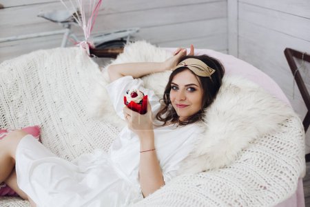 Téléchargez les photos : "Belle mariée en masque de sommeil et peignoir avec cupcake sur canapé confortable." - en image libre de droit