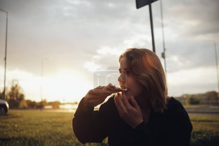 Téléchargez les photos : Blonde fille manger pizza assis dehors au coucher du soleil - en image libre de droit