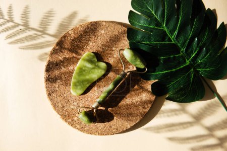Photo for "Jade Gua sha scraper and face roller massager on a cork round stand with a monstera leaf." - Royalty Free Image