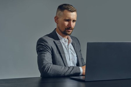 Téléchargez les photos : "Homme d'affaires courageux à la moustache élégante, vêtu d'un costume classique, travaille sur un ordinateur portable assis à une table au bureau, fond gris." - en image libre de droit