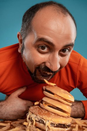 Téléchargez les photos : "Gros plan portrait d'un homme d'âge moyen barbu, vêtu d'un col roulé rouge, posant avec des hamburgers et des frites. Fond bleu. Restauration rapide." - en image libre de droit