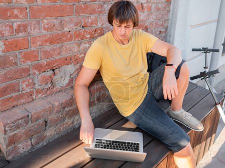 Téléchargez les photos : "Homme assis avec ordinateur portable et trottinette dans le café de la rue. Indépendante au travail. L'élève apprend à distance de l'extérieur. Style de vie moderne. Des vibrations estivales. Lieu de travail en plein air." - en image libre de droit