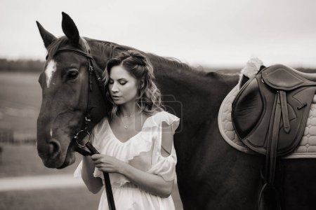 Foto de Una chica en un vestido blanco se para junto a un caballo marrón en un campo en verano. foto en blanco y negro - Imagen libre de derechos