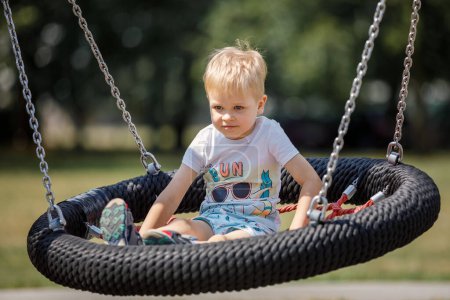 Photo for "Little blonde boy playing happily on large nest swing at city playground." - Royalty Free Image