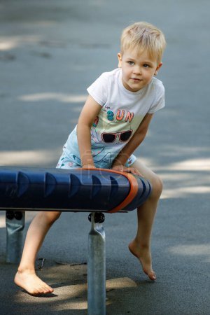 Photo for "Portrait of a little boy in an outdoor sports gym, a child performs gymnastic exercises barefoot on modern equipment" - Royalty Free Image