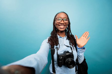Téléchargez les photos : "Happy smiling photographer with DSLR camera waving at audience while recording daily vlog video." - en image libre de droit