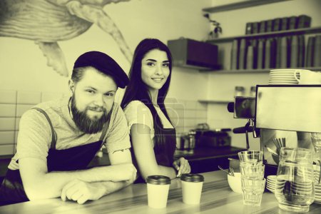 Téléchargez les photos : "Café Business Concept - Positif jeune homme barbu et belle belle dame couple barista attrayant dans tablier regardant la caméra tout en se tenant debout au comptoir du bar
" - en image libre de droit