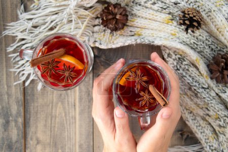 Photo for Female hands hold a glass of hot punch. mulled wine on a wooden table next to a warm knitted scarf. a warming drink in winter or autumn. Soft focus. view from above - Royalty Free Image