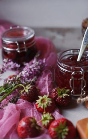 Photo for "Still life with srawberry jam in glass jars and fresh harvested berries with pink flowers on pink cloth. Summer breakfast concept." - Royalty Free Image