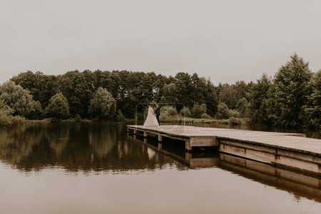 Téléchargez les photos : "Nouveaux mariés embrassant câlins sur pont jetée en bois sur le lac" - en image libre de droit