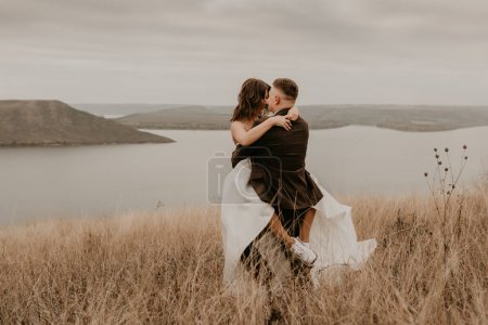 Téléchargez les photos : "couple amoureux mariage jeunes mariés en robe voile chaussures de sport et costume embrasser tourbillon sur l'herbe haute dans le champ sur la montagne au-dessus de la rivière" - en image libre de droit