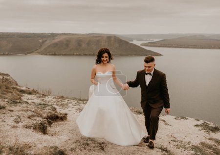 Téléchargez les photos : "mariée en robe blanche long voile et marié en costume marcher en été automne sur la montagne au-dessus de la rivière" - en image libre de droit
