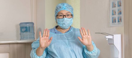 Photo for "banner with medical worker in a disposable medical suit, a medical cap and a mask with glasses, shows the treated hands with a disinfectant solution next to the elbow dispenser." - Royalty Free Image