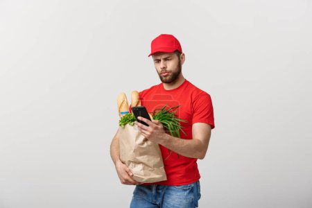 Téléchargez les photos : "Concept de livraison : beau livreur d'épicerie caucasien en uniforme rouge avec boîte d'épicerie avec fruits et légumes frais
" - en image libre de droit