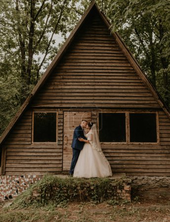Foto de "Recién casados abrazándose de pie cerca de la pequeña casa triangular de madera" - Imagen libre de derechos