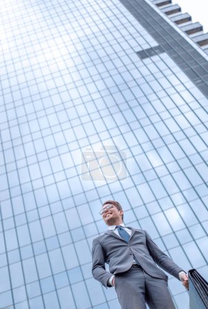 Téléchargez les photos : Homme d'affaires avec mallette debout sur l'herbe près du grand immeuble de bureaux - en image libre de droit