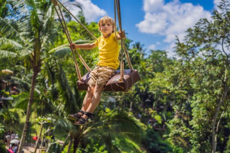 Téléchargez les photos : Jeune garçon se balançant dans la jungle de la forêt tropicale de Bali, Indonésie. Balancez sous les tropiques. Balançoires tendance de Bali. Voyager avec des enfants concept. Que faire avec les enfants ? Endroit adapté aux enfants - en image libre de droit