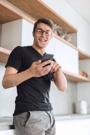 Téléchargez les photos : Beau mec avec une tasse de café et un smartphone debout dans la cuisine - en image libre de droit