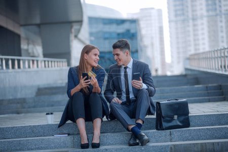 Foto de Joven pareja de negocios con smartphones sentados cerca del edificio de oficinas. - Imagen libre de derechos