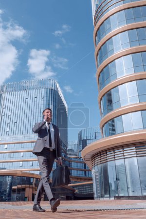 Photo for Businessman talking on a smartphone, walking along a city street - Royalty Free Image