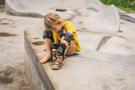 Photo for "Boy puts on knee pads and armbands before training skate board" - Royalty Free Image