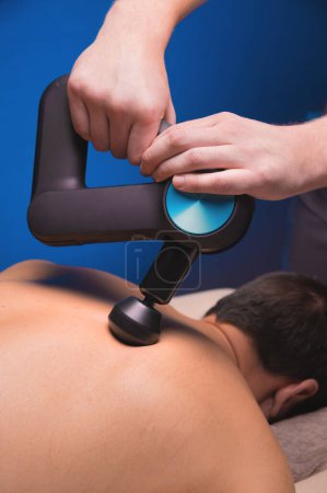 Photo for "Medium shot of caucasian professional male massage therapist getting back muscles with massage gun impact tool of muscular athlete, in spa treatments, lying on massage table." - Royalty Free Image