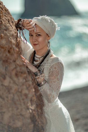 Foto de "Middle aged woman looks good with blond hair, boho style in white long dress on the beach decorations on her neck and arms." - Imagen libre de derechos