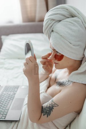 Photo for "Tver, Russia-august 2, 2021 A woman in tattoos after a shower is lying on the couch with a laptop and an eye mask at the same time and looks in the mirror." - Royalty Free Image
