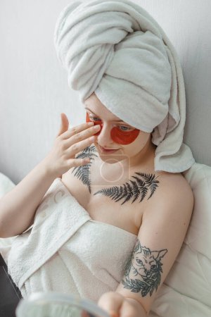 Photo for "Tver, Russia-august 2, 2021 A woman in tattoos after a shower is lying on the couch with a laptop and an eye mask at the same time and looks in the mirror." - Royalty Free Image