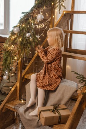 Photo for A little blonde girl is sitting on a wooden staircase in a Scandinavian interior decorated. - Royalty Free Image