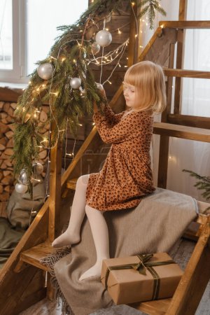 Foto de A little blonde girl is sitting on a wooden staircase in a Scandinavian interior decorated. - Imagen libre de derechos