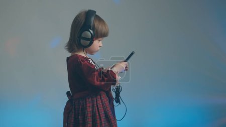 Téléchargez les photos : Little girl in big headphones and vintage dress is listening to music on phone - en image libre de droit