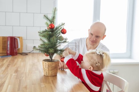 Foto de Child with cochlear implant hearing aid having fun with father and small christmas tree - diversity and deafness treatment and medical innovative technologies - Imagen libre de derechos