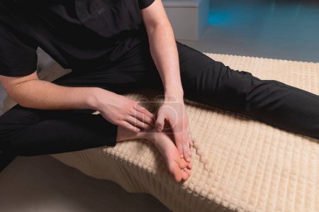 Téléchargez les photos : Un homme assis sur une table de massage fait l'auto-massage de ses pieds avec ses propres mains - en image libre de droit
