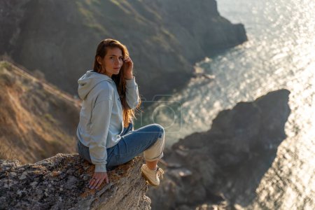 Téléchargez les photos : "Femme touriste profitant du coucher de soleil sur le paysage de montagne de la mer. Assis à l'extérieur sur un rocher au-dessus de la mer. Elle porte un jean et une capuche bleue. Mode de vie sain, harmonie et méditation" - en image libre de droit