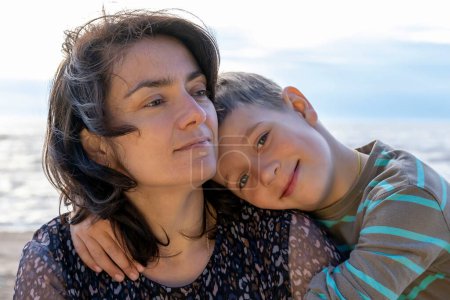 Téléchargez les photos : Cute happy boy hugs his mom by the sea. looking into the camera - en image libre de droit