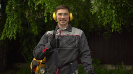 Photo for "A young man from the special services mows the lawn with a petrol trimmer. A man in glasses and headphones looks at the camera, smiles and shows a thumbs up." - Royalty Free Image