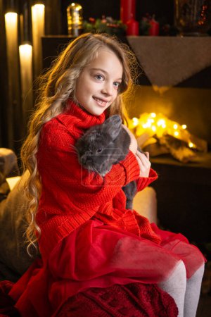 Photo for A little blonde girl with a gray rabbit in her arms next to a Christmas tree decorated with garlands - Royalty Free Image