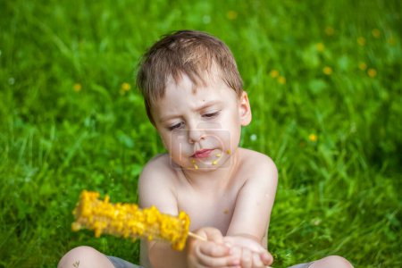 Foto de "Un lindo chico rubio come maíz en verano, sentado en la orilla del río sobre la jugosa hierba. Expresión facial divertida." - Imagen libre de derechos