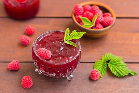 "fresh raspberry jam in a glass jar on a wooden table, next to fresh raspberries. concept of homemade jam, preserves for winter, selective focus"