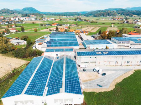 Foto de Paneles solares instalados en un techo de un gran edificio industrial o un almacén. Edificio industrial en el campo de Eslovenia con casas residenciales en el fondo. Foto de alta calidad - Imagen libre de derechos