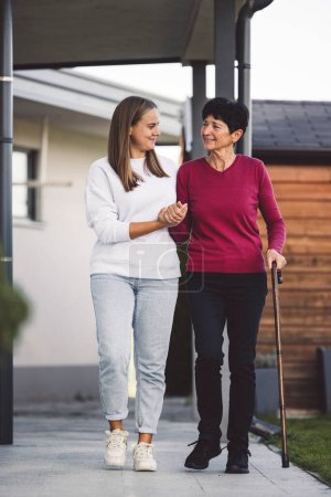 Photo for Young caucasian woman a nurse, on a home visit taking care of a senior lady. Nurse helping a senior women with every day chores, helping her to walk and move around the house. - Royalty Free Image
